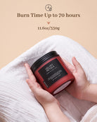 Red candle with black lid held by hands on a beige background, featuring text about burn time and weight.