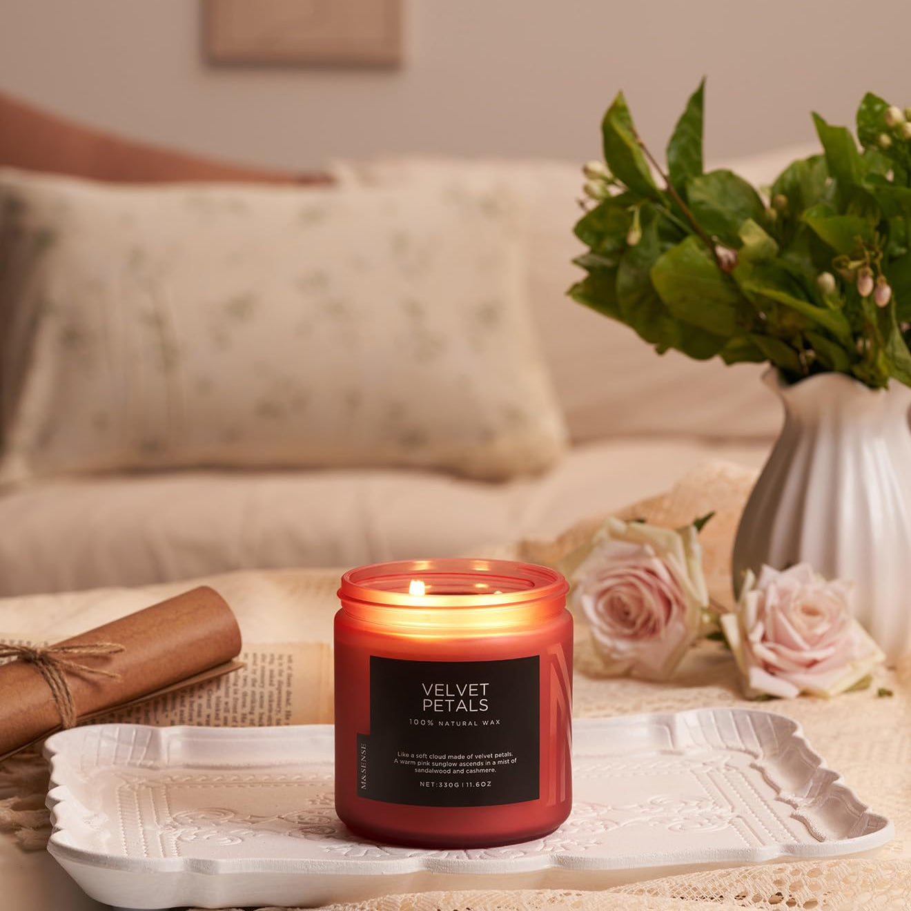 Candle labeled 'Velvet Petals' on a table with a vase of flowers and a blanket in the background