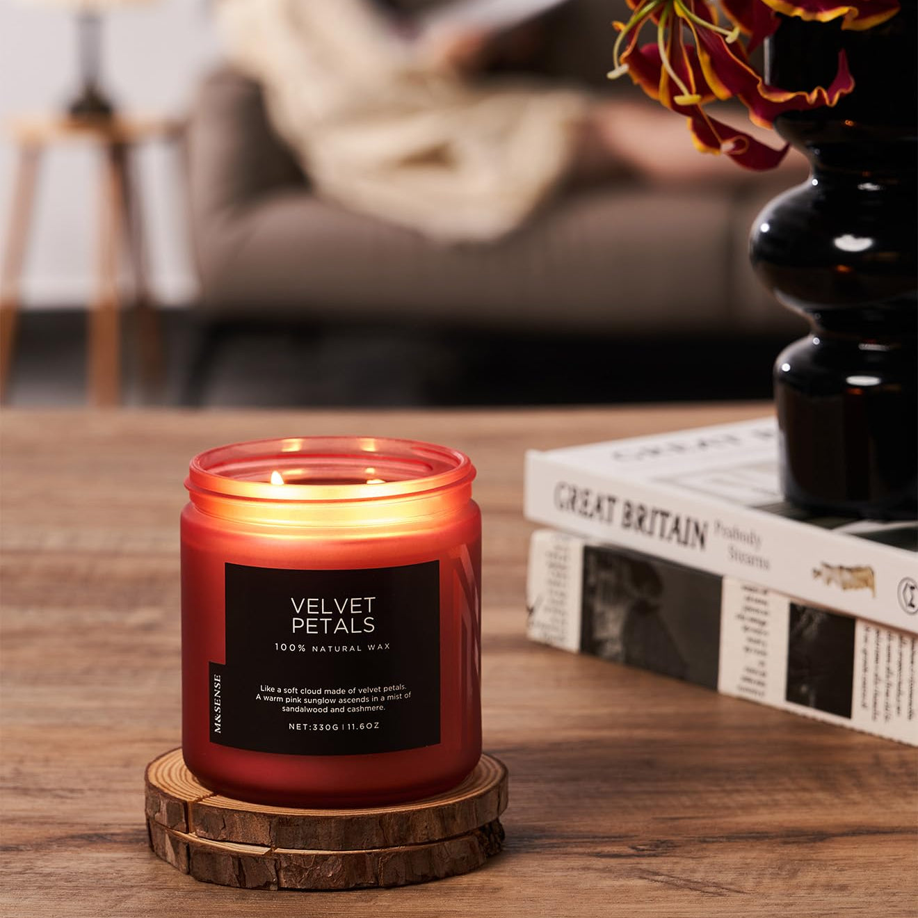 Candle labeled 'Velvet Petals' on a wooden coaster with a blurred background of a person reading.