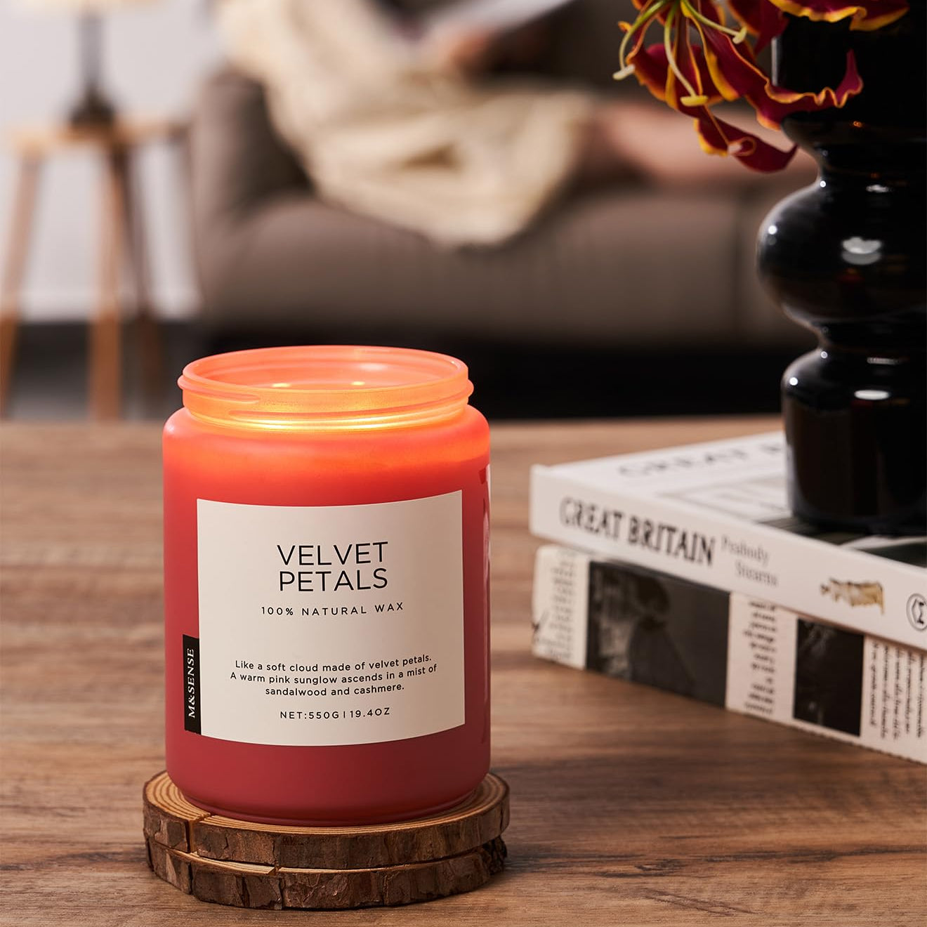 Candle labeled 'Velvet Petals' on a wooden coaster with a blurred background of a person reading a book.
