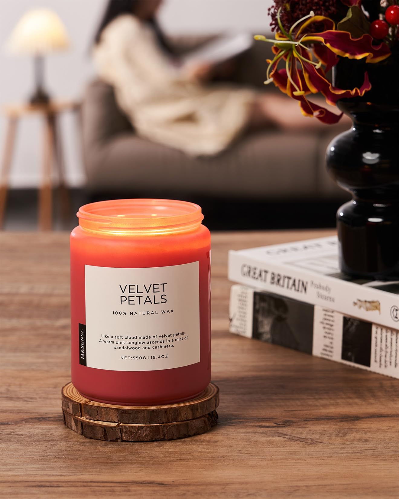 Candle labeled 'Velvet Petals' on a wooden coaster with a blurred background of a person reading a book.