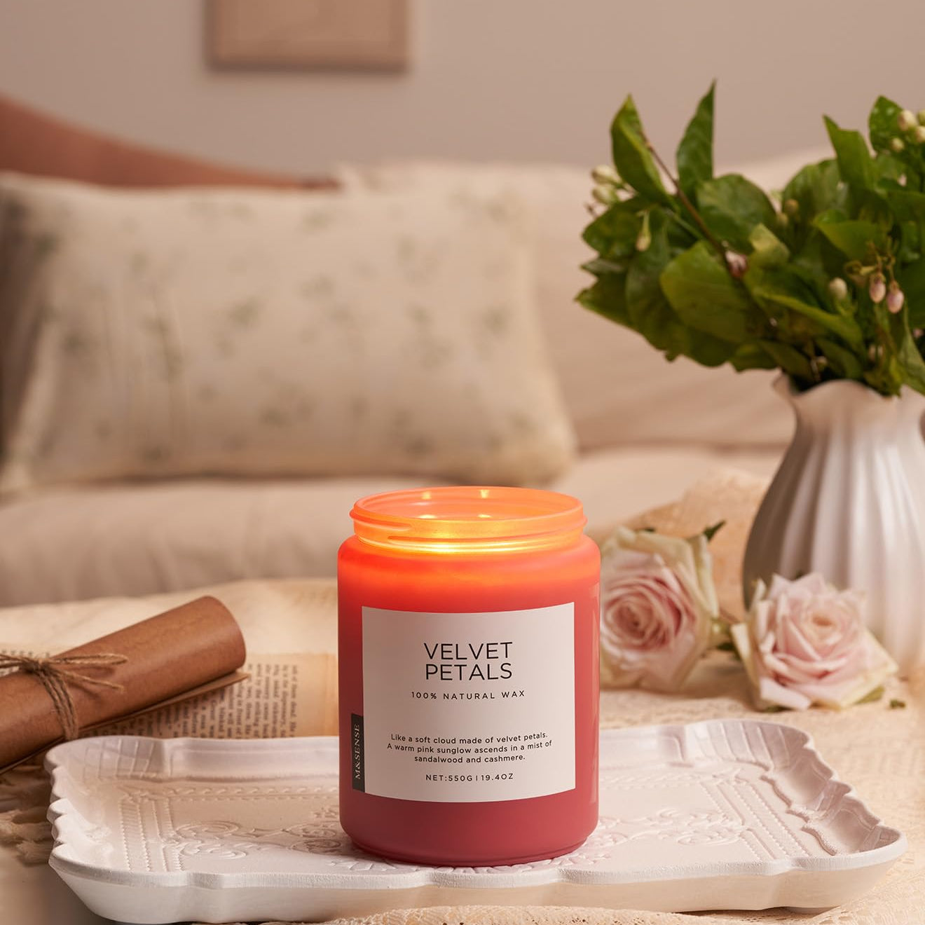Pink candle labeled 'Velvet Petals' on a tray with a vase of flowers and a couch in the background.