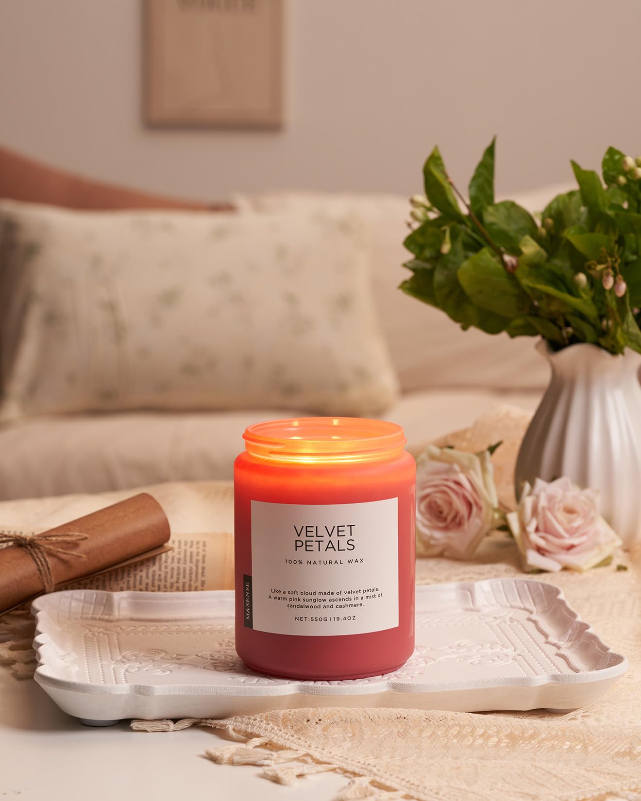 Pink candle labeled 'Velvet Petals' on a tray with a vase of flowers and a couch in the background.