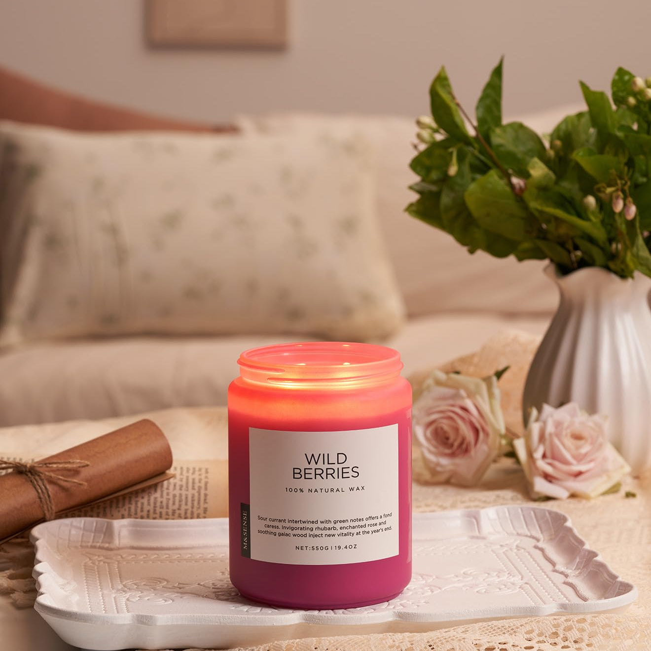Pink candle labeled 'Wild Berries' on a table with a vase of flowers and a couch in the background.
