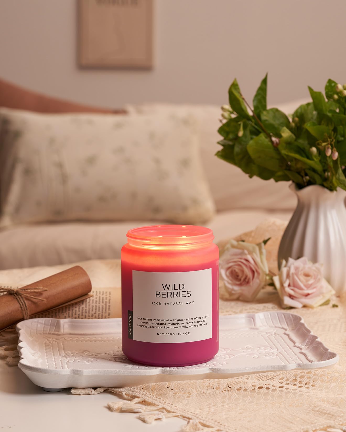 Pink candle labeled 'Wild Berries' on a table with a vase of flowers and a couch in the background.