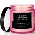 A pink candle with a label reading 'Cherry Blossom' and '100% Natural Wax'. The candle is placed in a cylindrical pink container with a black lid, which is partially open.
