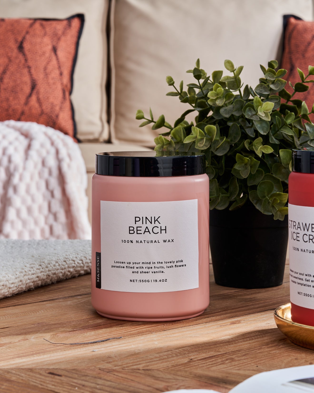 Pink Beach scented candle on a wooden table with a green plant, against a sofa background.