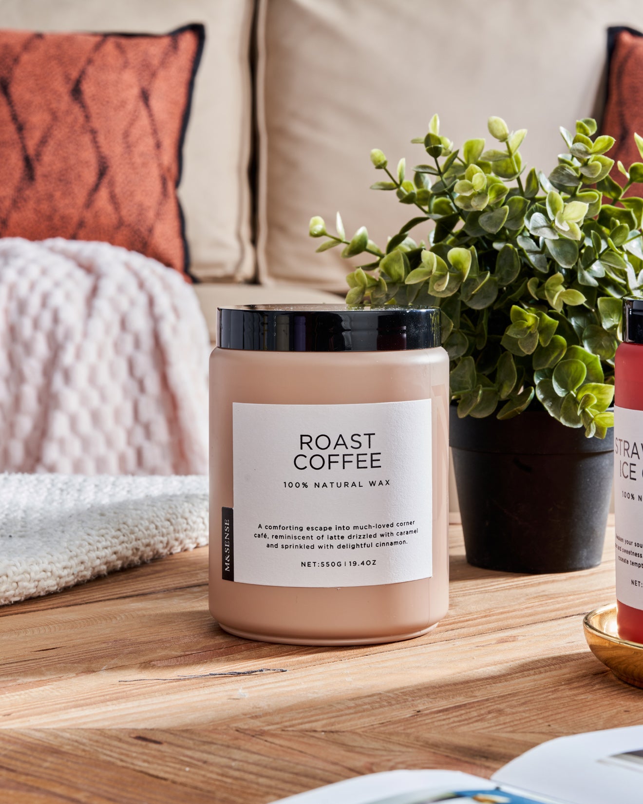 Pink Roast Coffee scented candle on a wooden table with a green plant, against a sofa background.