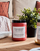 Red Strawberry Ice Cream scented candle on a wooden table with a green plant, against a sofa background.