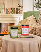 Two scented candles on a draped round coffee table, in a living room with sofa and bookshelf.