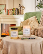 Two scented candles on a draped round coffee table, in a living room with sofa and bookshelf.