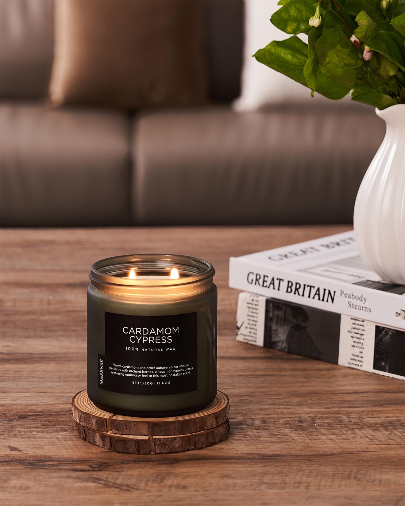 Candle labeled 'Cardamom Cypress' on a wooden coaster with a blurred sofa and plant in the background.