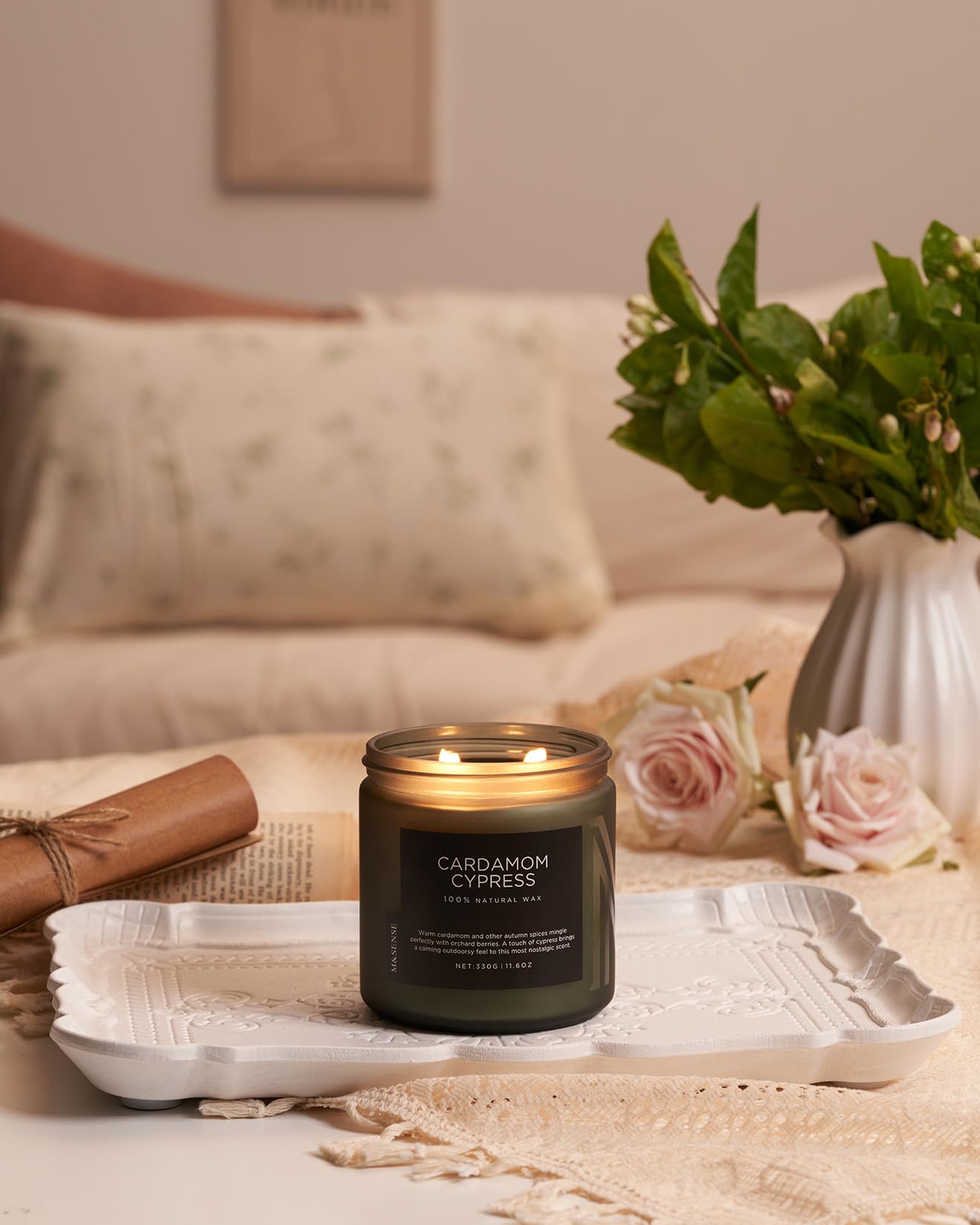 Candle labeled 'Cardamom Cypress' on a tray with flowers and a vase in the background