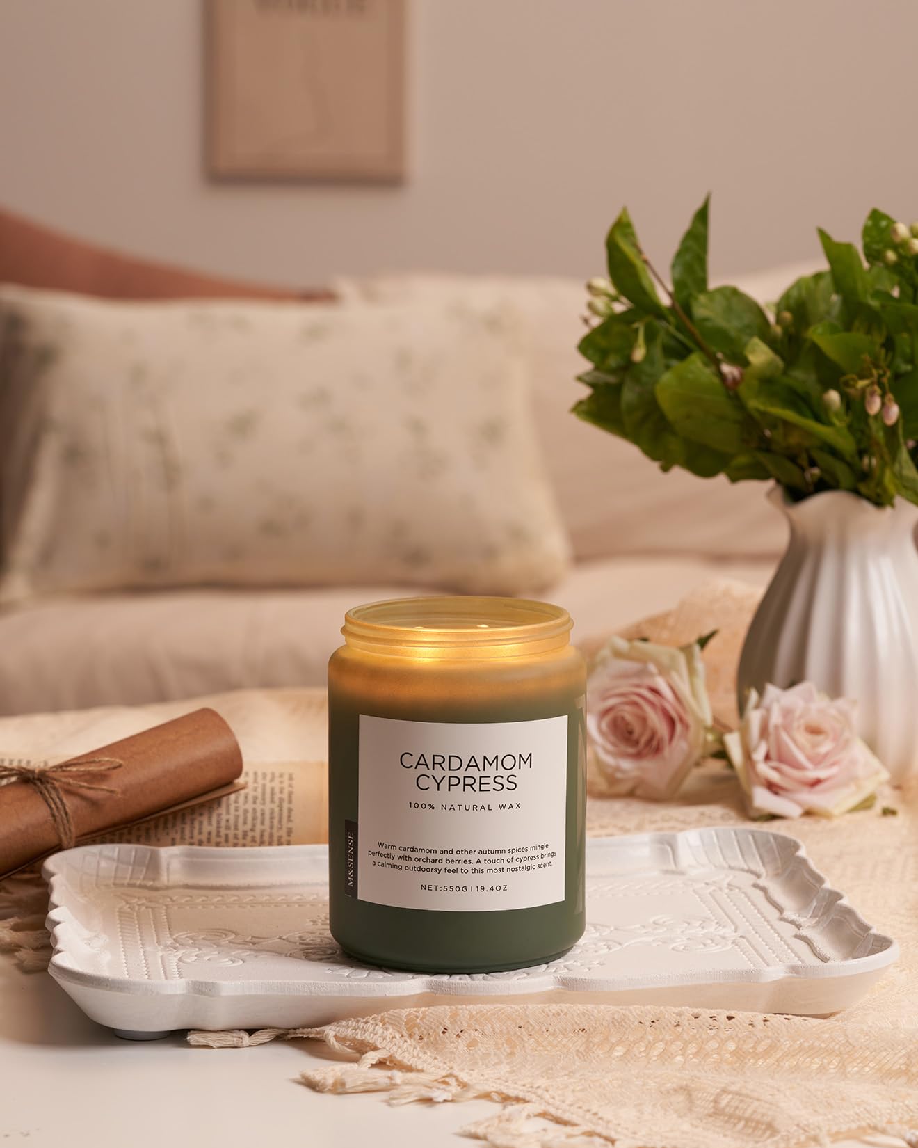 Candle labeled 'Cardamom Cypress' on a table with flowers and a vase in the background