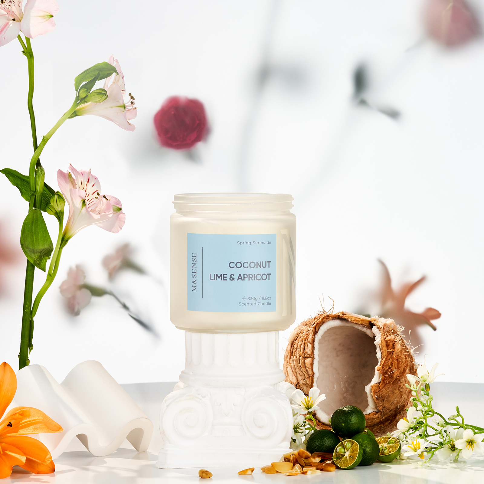Candle labeled 'Coconut Lime & Apricot' with a coconut shell and fruits on a white surface with flowers in the background.