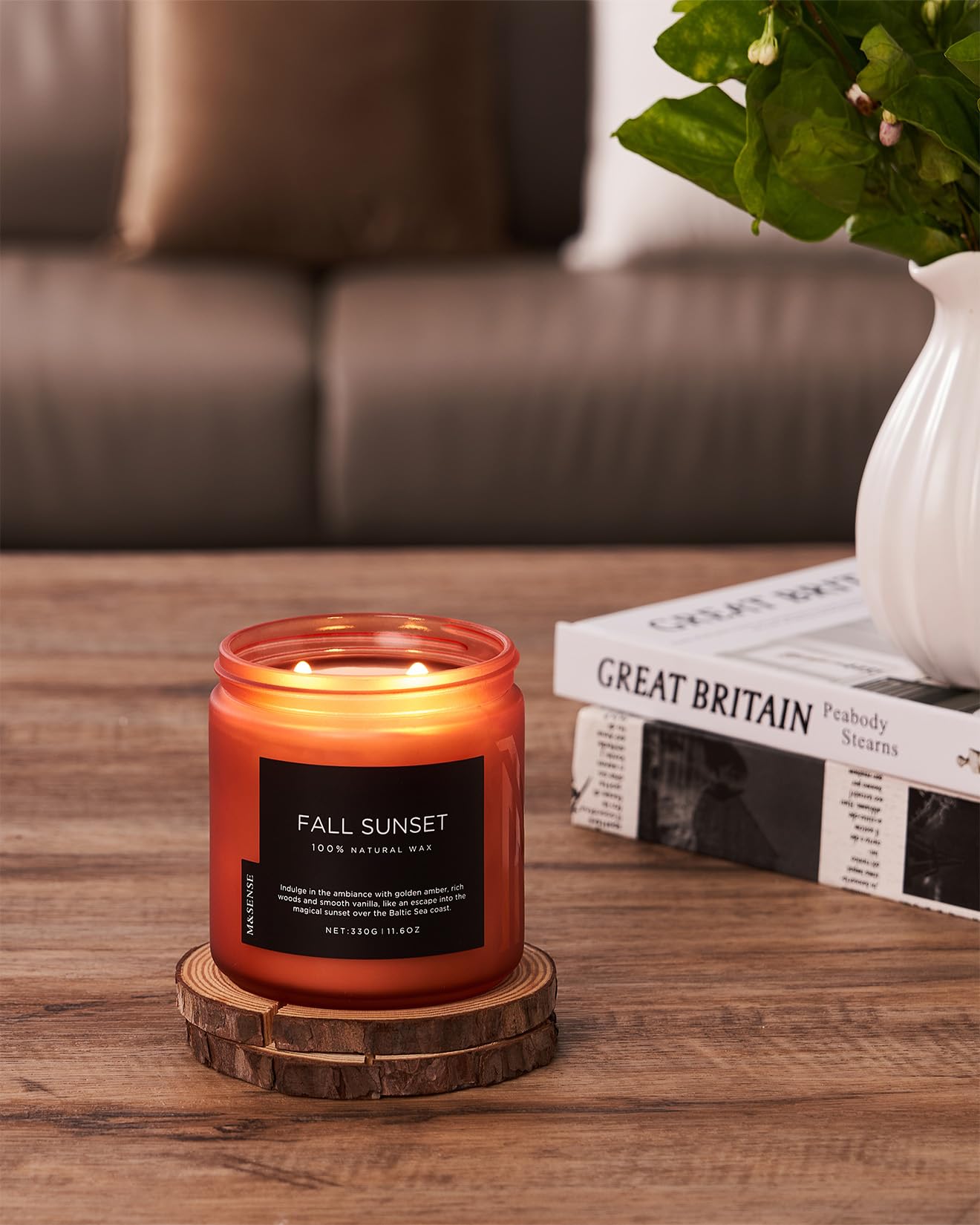 Candle labeled 'Fall Sunset' on a wooden coaster with a blurred background of a couch and books.