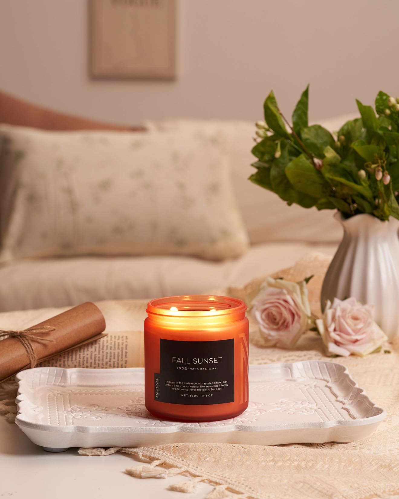 Candle labeled 'Fall Sunset' on a table with flowers and a vase in the background