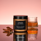 Candle labeled 'Fireside Bourbon' with a glass of whiskey and cinnamon sticks on a reflective surface.