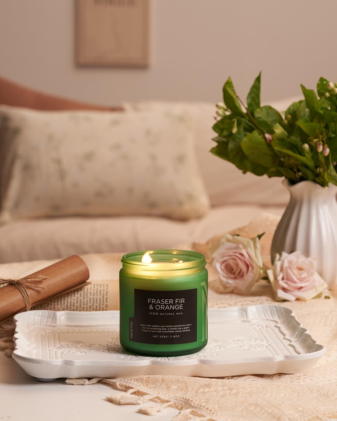 Green candle labeled 'Fraser Fir & Orange' on a tray with flowers and a vase in the background.