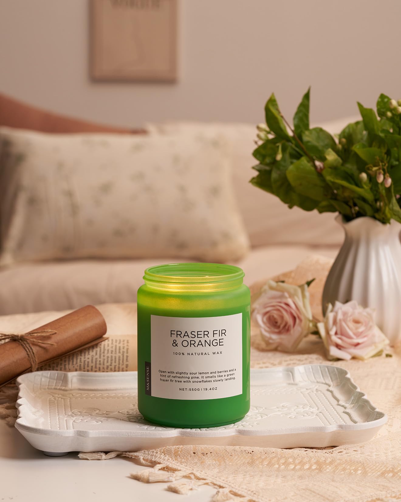 Green candle labeled 'Fraser Fir & Orange' on a tray with flowers and a vase in the background.