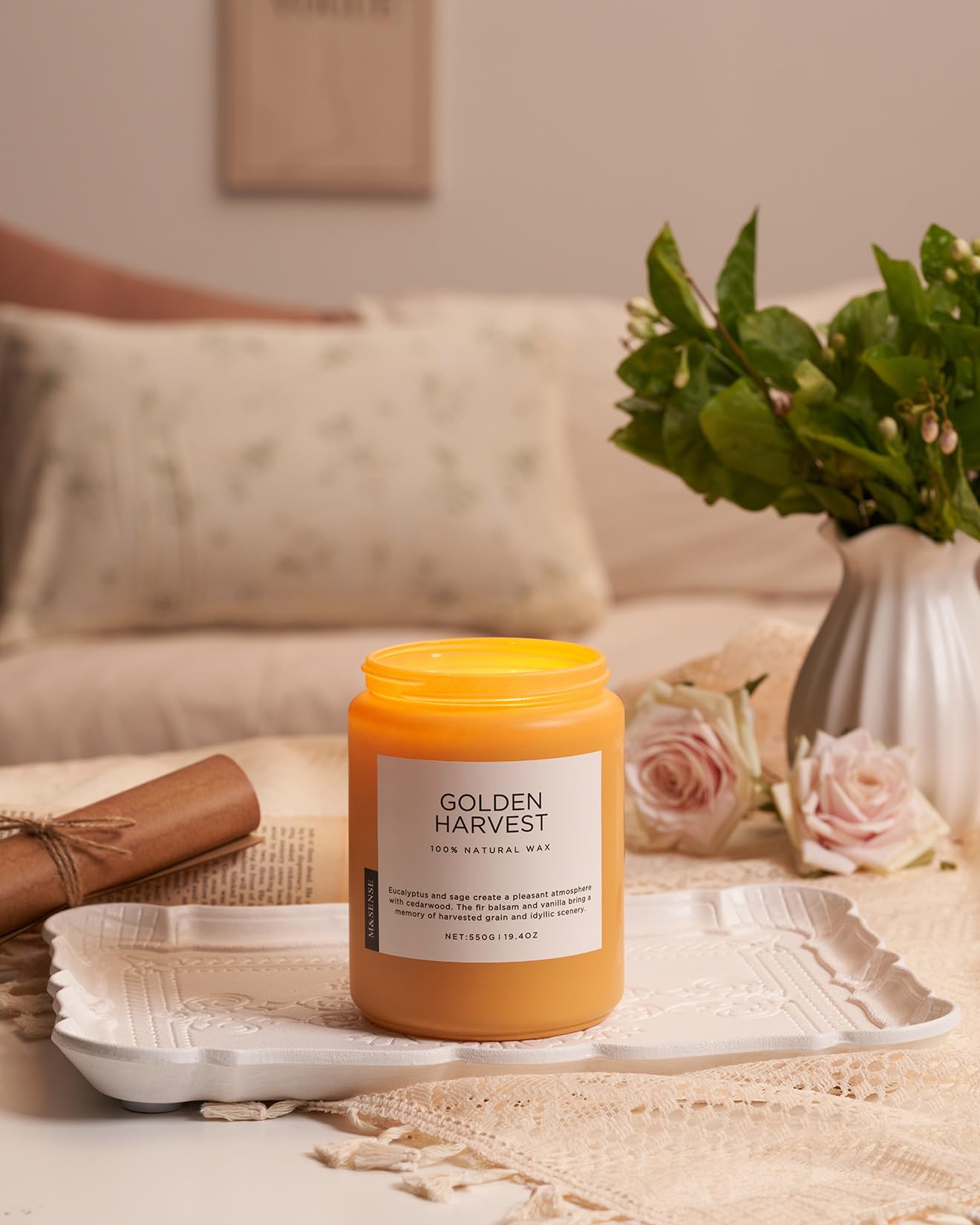 Candle labeled 'Golden Harvest' on a table with flowers and a couch in the background