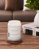 Candle labeled 'White Lazy' on a wooden coaster with a blurred sofa and plant in the background