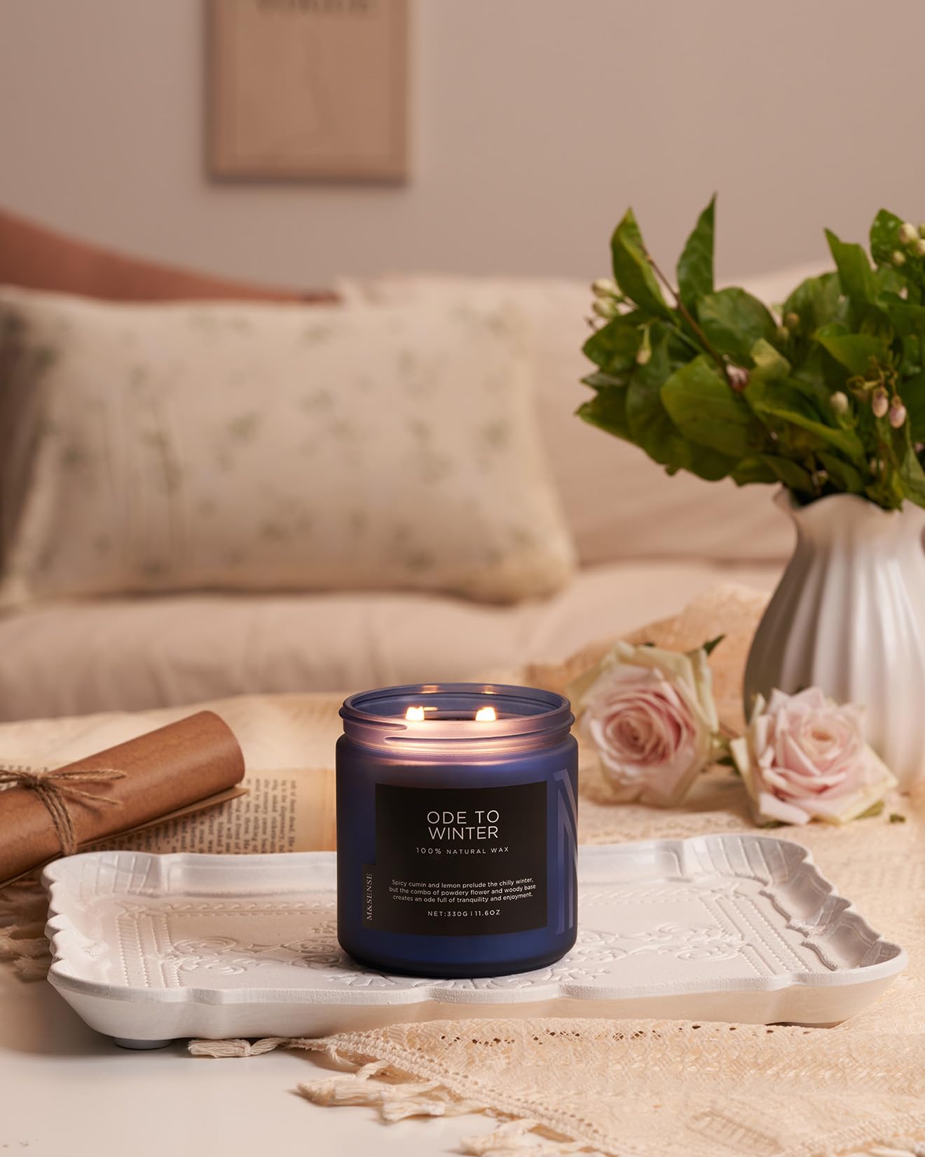 Candle labeled 'Ode to Winter' on a decorative tray with flowers and pillows in the background