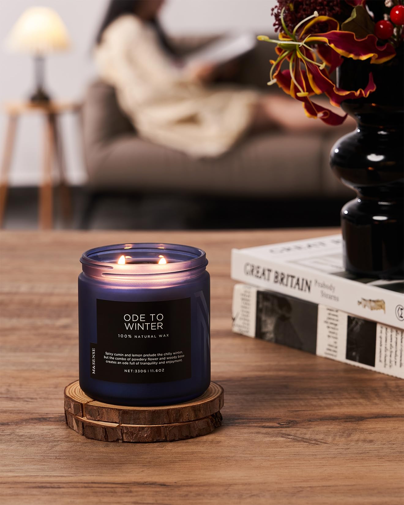 Candle labeled 'Ode to Winter' on a wooden coaster with a blurred background of a person reading.