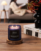 Candle labeled 'Ode to Winter' on a wooden coaster with a blurred background of a person reading.