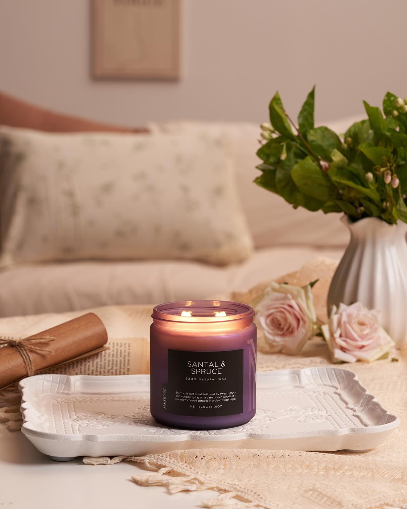 Scented candle labeled 'Santal & Spruce' on a tray with flowers and a vase in the background.