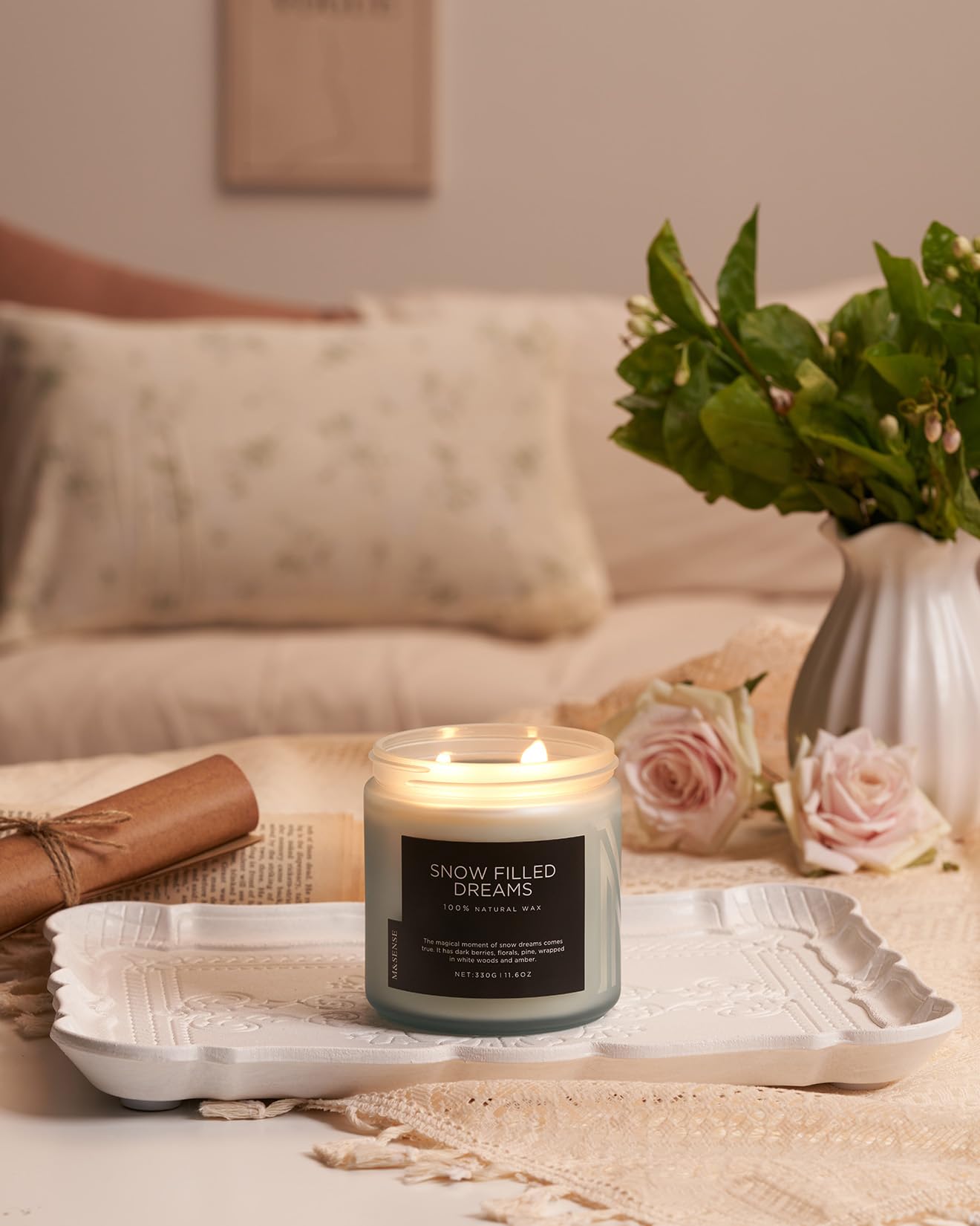 Candle labeled 'Snow Filled Dreams' on a table with a vase of flowers and a blanket in the background.