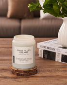 Candle labeled 'Snow Filled Dreams' on a wooden coaster with a vase and books in the background.