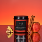 Spiced Apple candle with ginger, apple, and cinnamon sticks on a reflective surface with an orange gradient background.