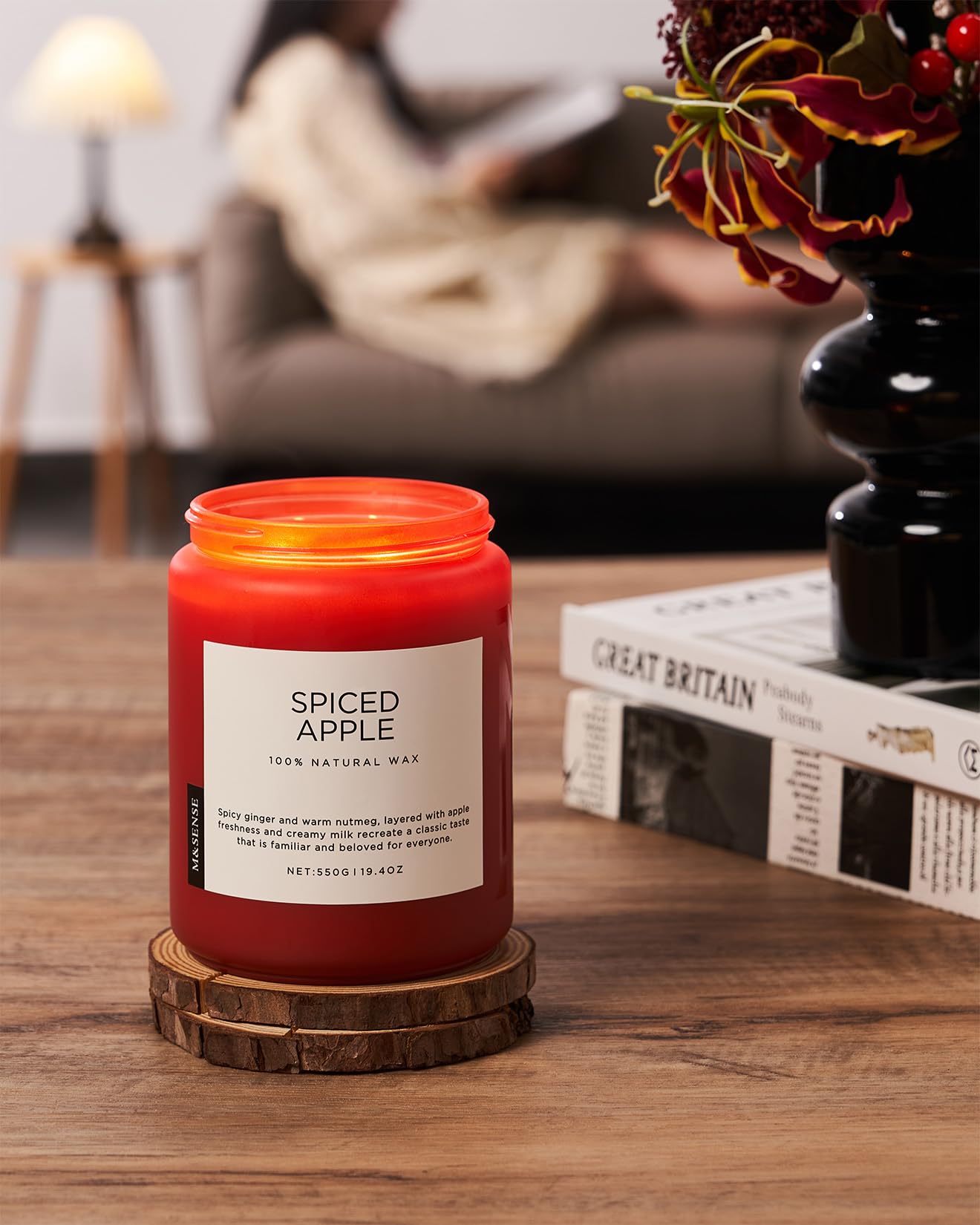 Red candle labeled 'Spiced Apple' on a wooden coaster with a blurred background of a person reading a book.